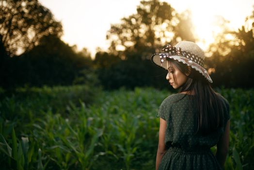 A woman wearing a sun hat stands in a lush green field at sunset, surrounded by nature.