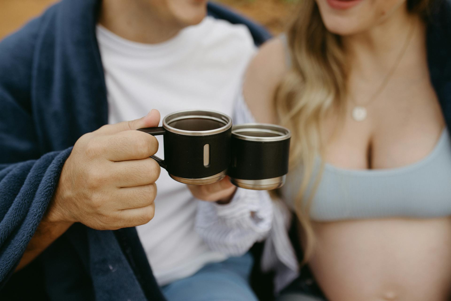 Close-up of a couple clinking insulated mugs, wrapped in blankets outdoors.