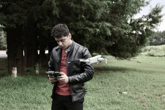 A young man with a controller and drone in a green park, wearing a leather jacket.