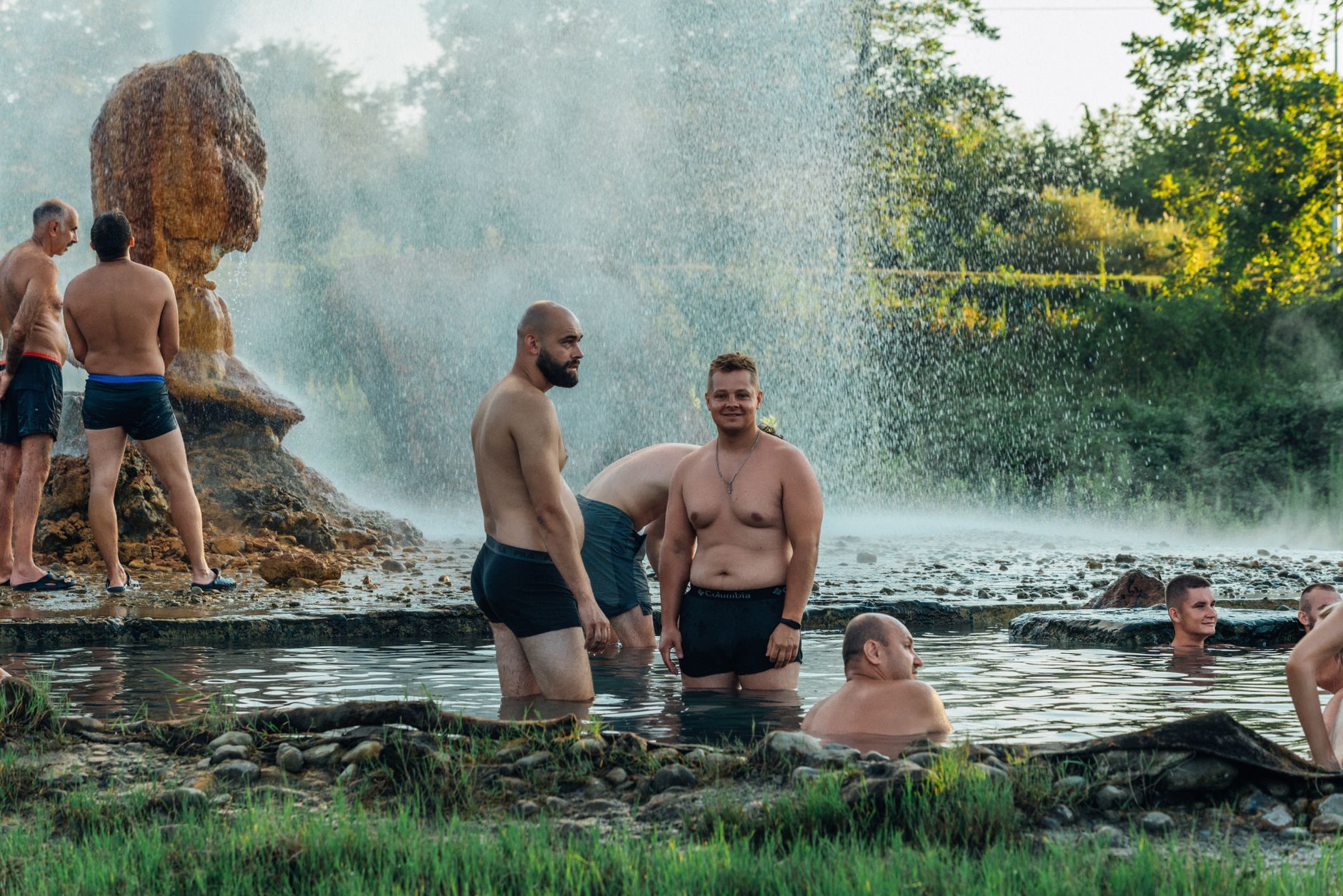 Group of men enjoying natural hot springs outdoors. Steam rises, creating a tranquil scene.