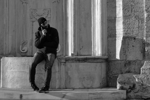 Black and white image of a man using a smartphone, leaning on a historic stone architecture.