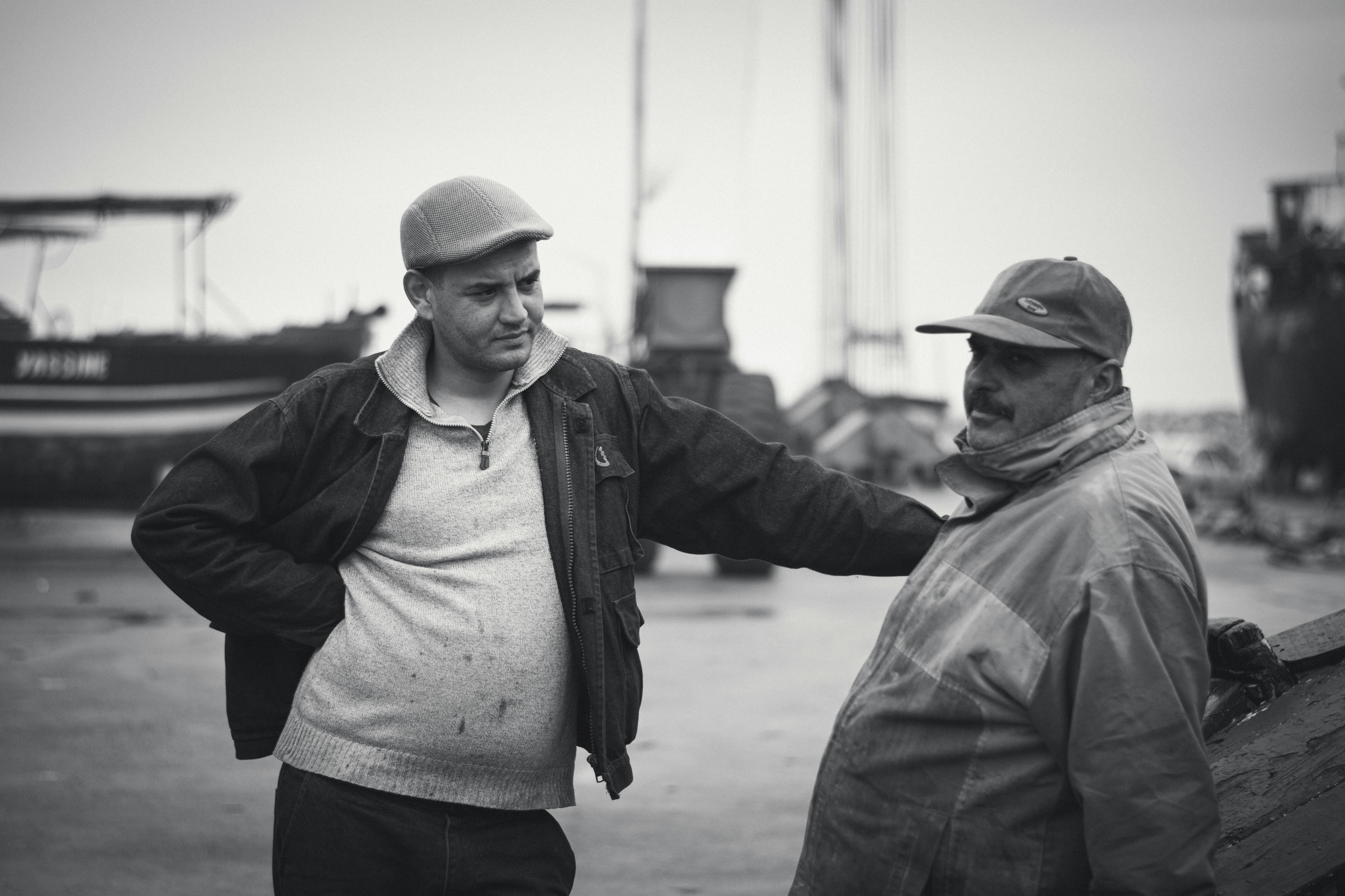 Two fishermen sharing a moment at Tabarka harbor in Tunisia, captured in black and white.