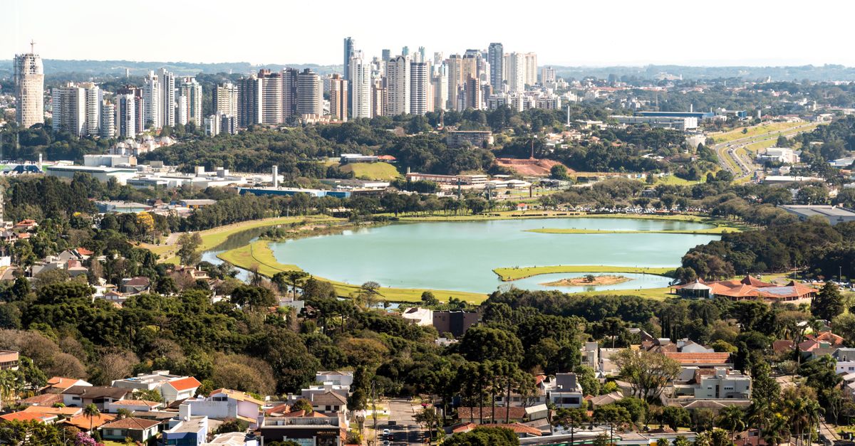 Vista aérea do skyline de Curitiba com o lago do Parque Barigui ao fundo, mostrando a integração entre natureza e cidade