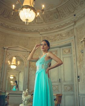 Stylish woman in vintage gown posing in an opulent room with antique decor and chandeliers.