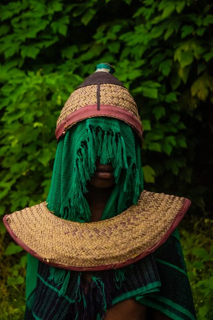 A person wearing traditional woven attire and hat amidst lush green foliage.