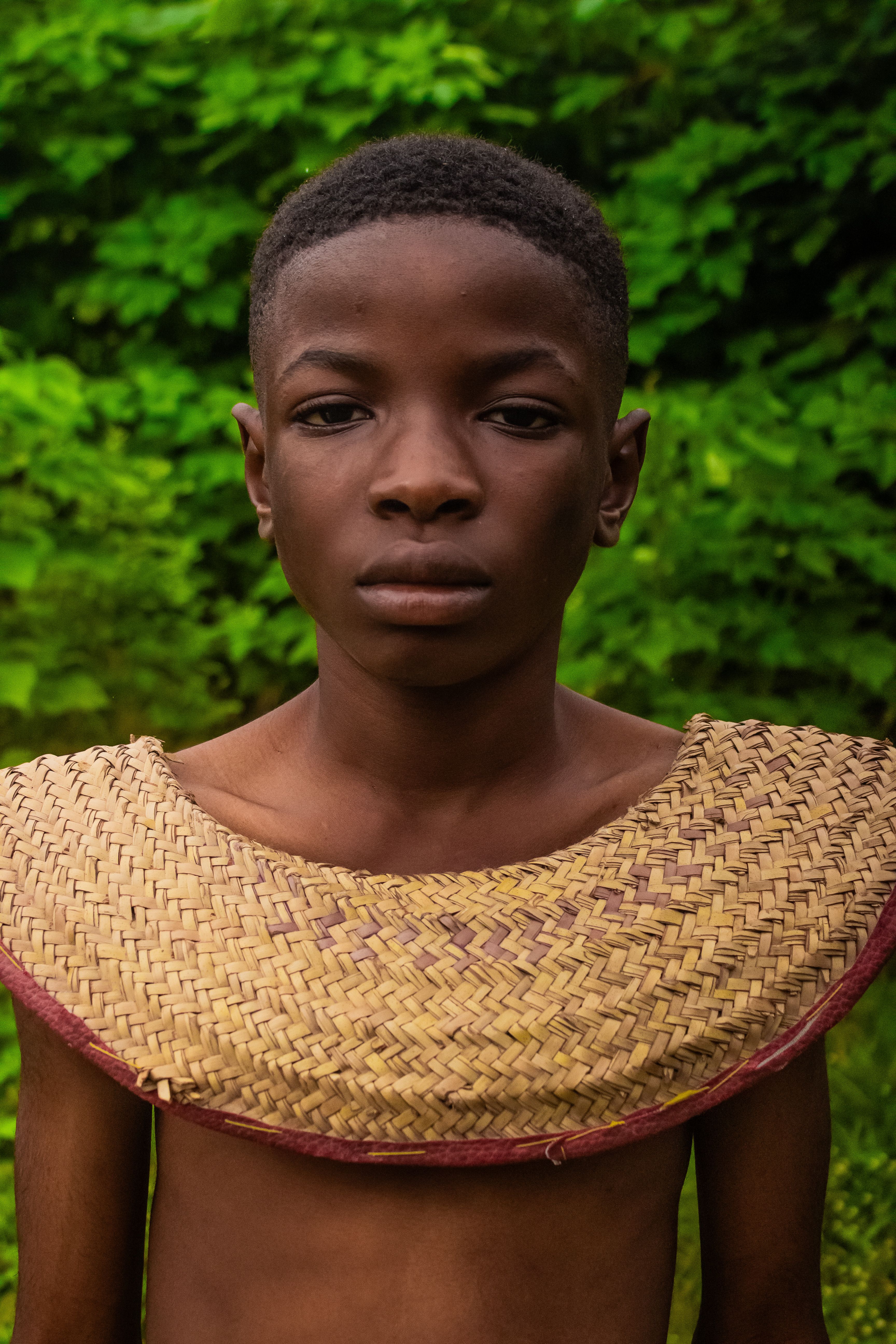 Close-up portrait of a young boy wearing a traditional woven garment, standing outdoors with a green leafy background.