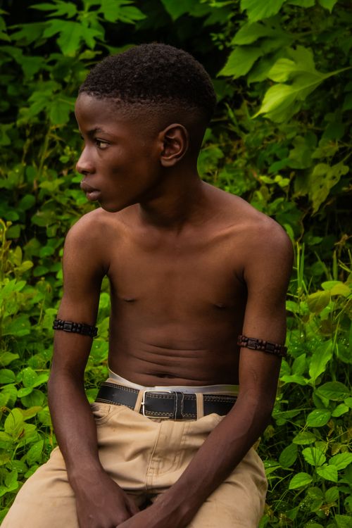 Free Portrait of a young boy in nature wearing arm bracelets, sitting among lush green foliage. Stock Photo