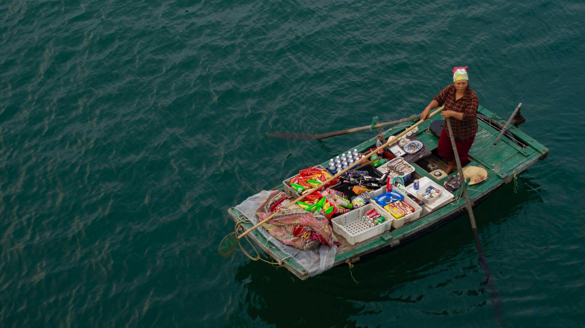 Aerial view of an Asian woman rowing a merchant boat filled with goods on a tranquil river.