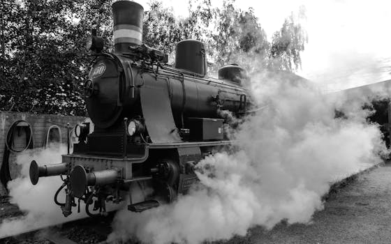 Black and white image of a vintage steam locomotive releasing steam clouds outdoors.