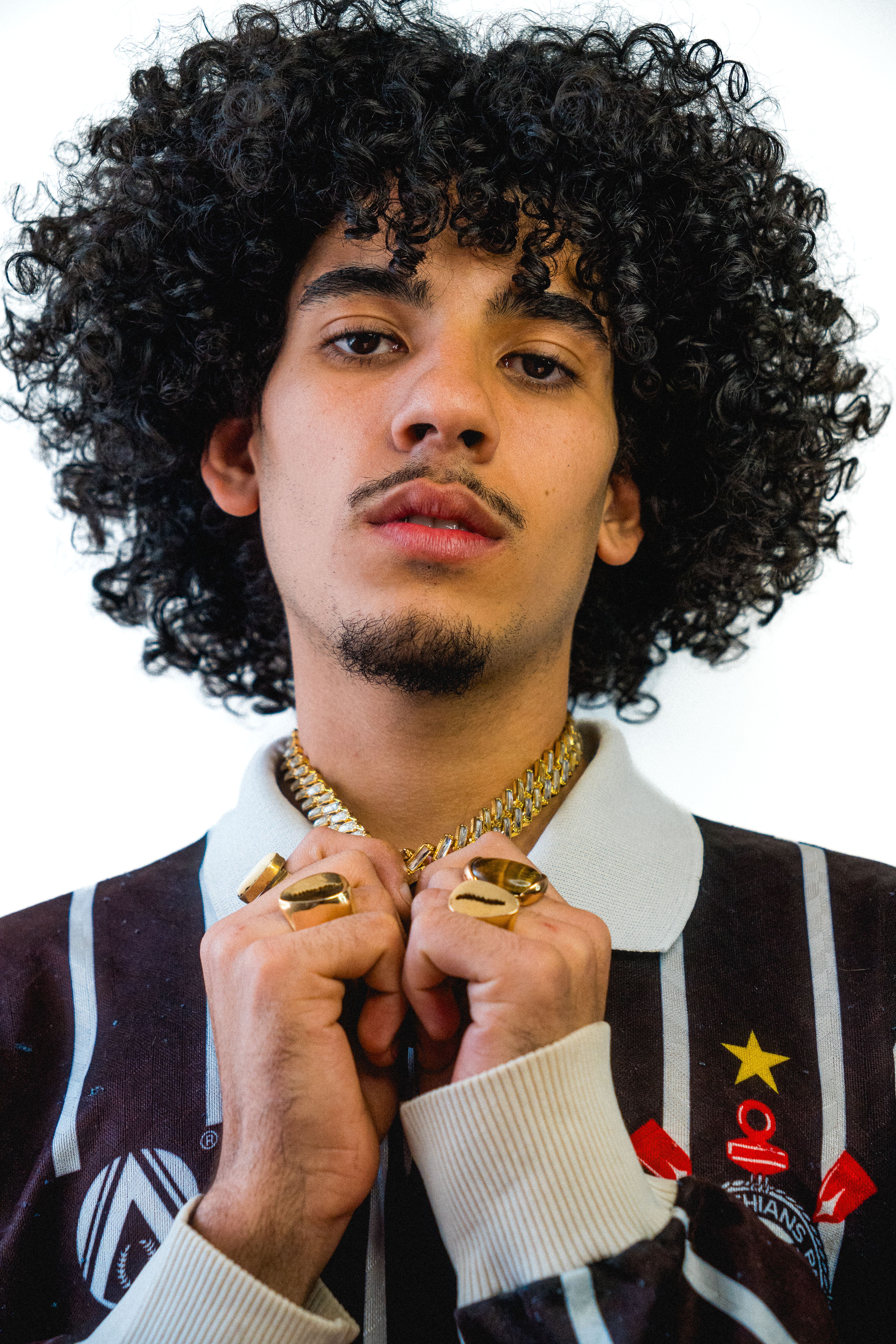 Portrait of a stylish young man with curly hair, wearing gold jewelry and a sporty jacket.