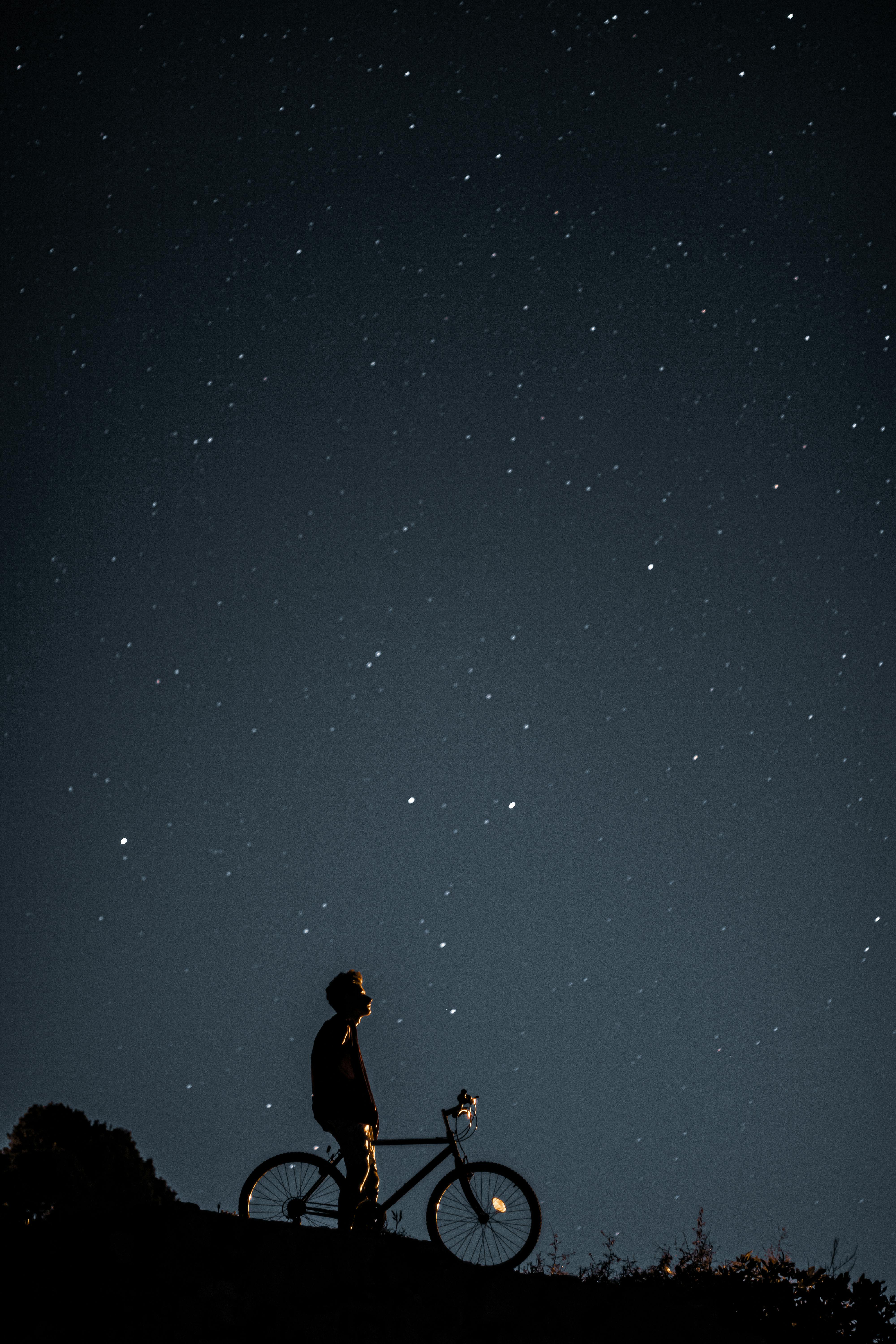 Silhouette of a person with a bicycle gazing at a star-filled night sky, evoking exploration.