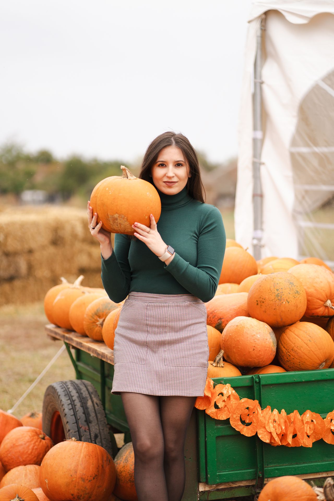 Woman in a dark green turtleneck and plaid mini skirt with black tights holding a pumpkin beside a farm cart