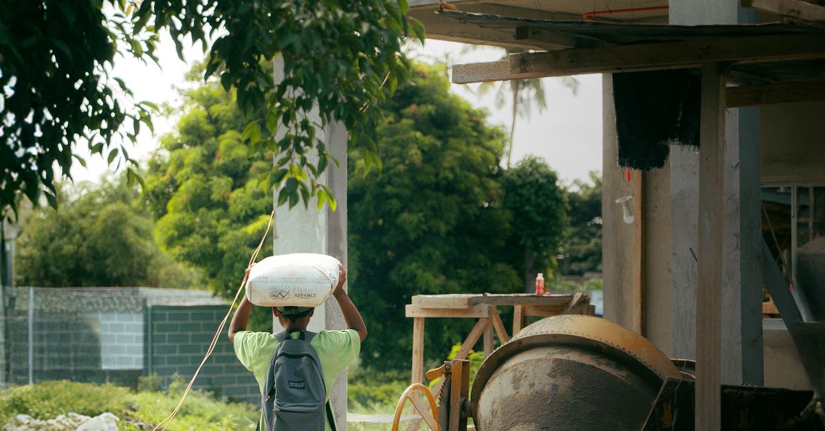 Photo by Kenneth Surillo A worker carrying cement at a construction site surrounded by greenery.
