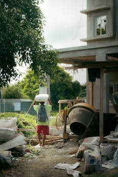A worker carrying cement at a construction site surrounded by greenery.