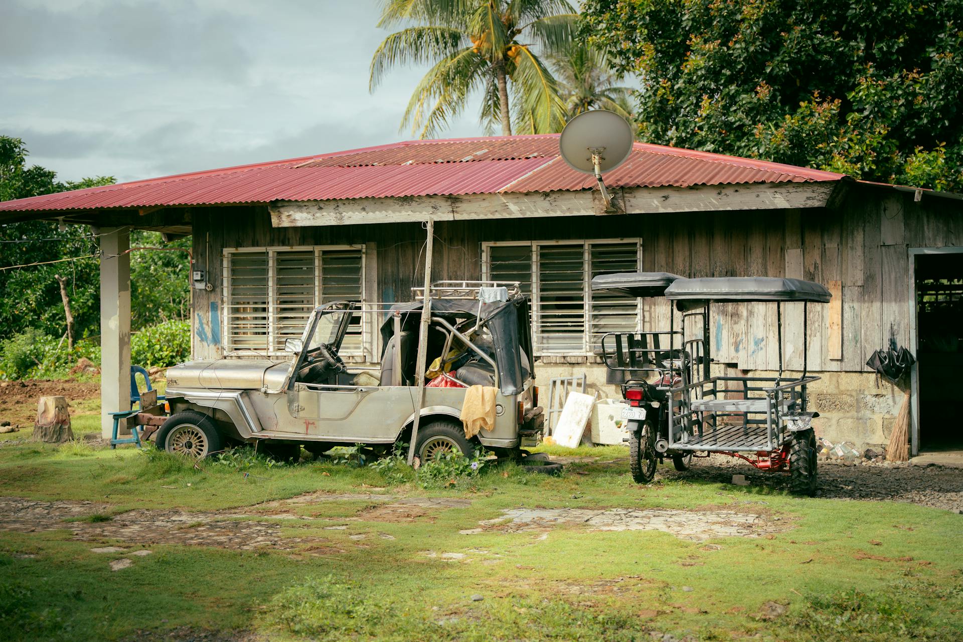 A wooden house with an old vehicle and tricycle in a rural setting.