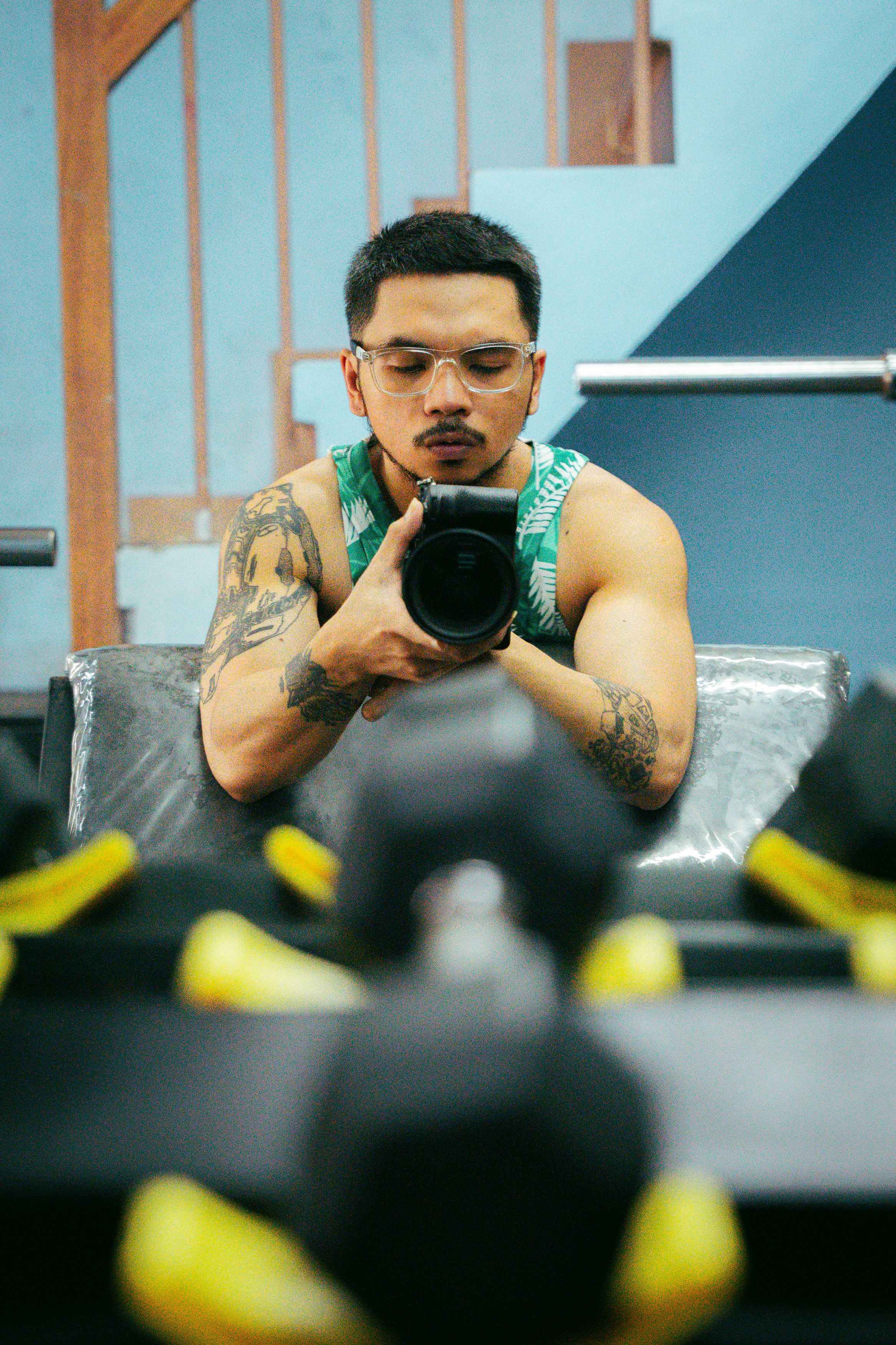 Photographer with tattoos taking a mirror selfie in gym surrounded by dumbbells.
