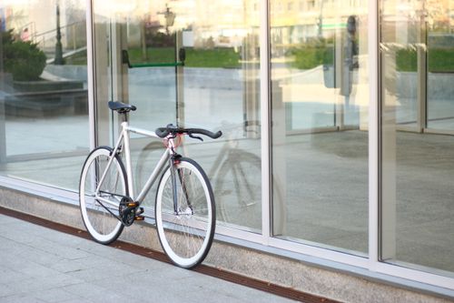 Free A sleek white bicycle rests against a glass building wall, reflecting urban style. Stock Photo