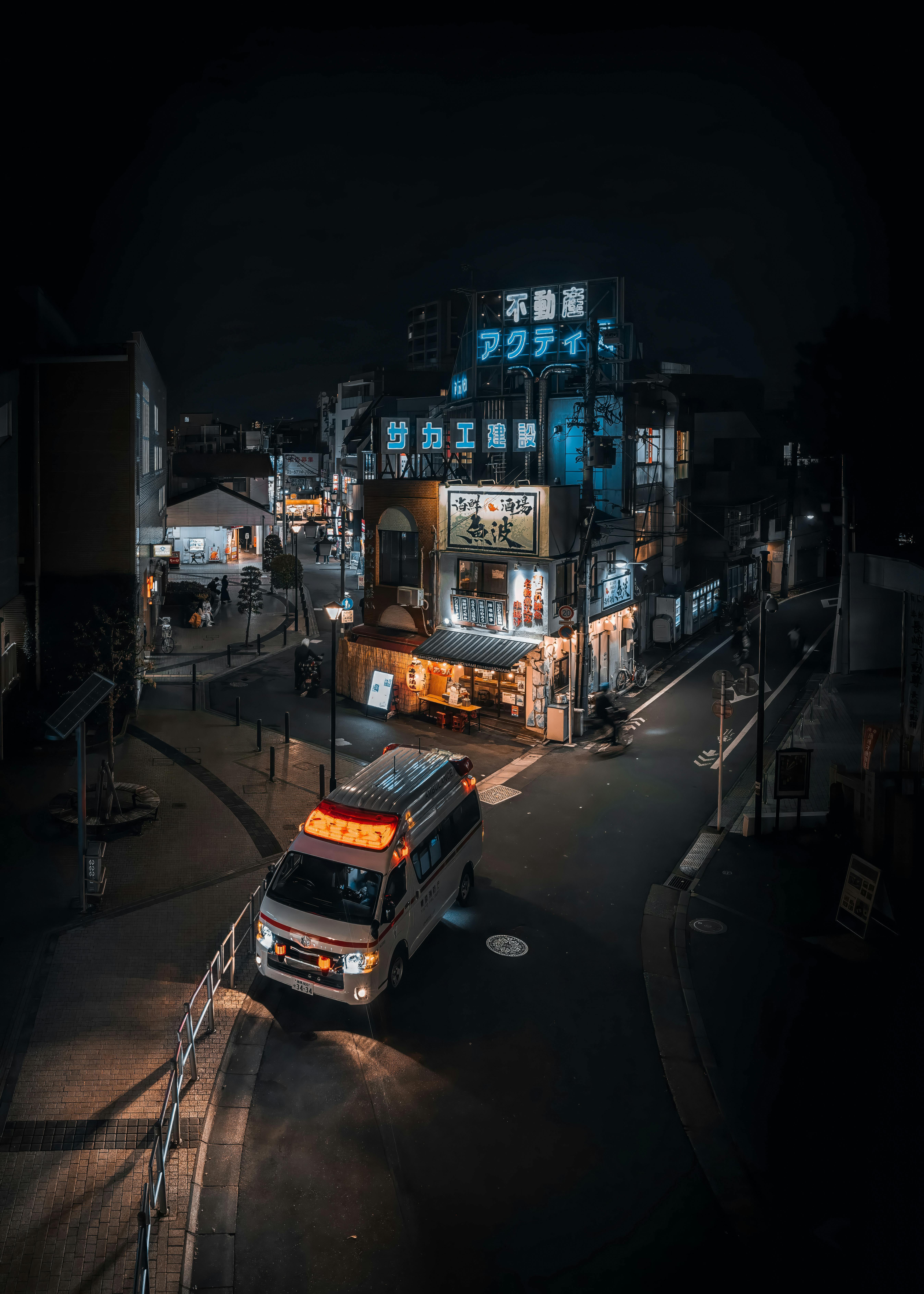 Street view of Tokyo at night featuring a neon-lit izakaya and an ambulance in motion.
