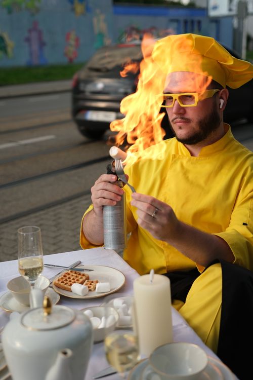 Free A chef in yellow attire uses a culinary torch to create a dramatic display with food outdoors. Stock Photo