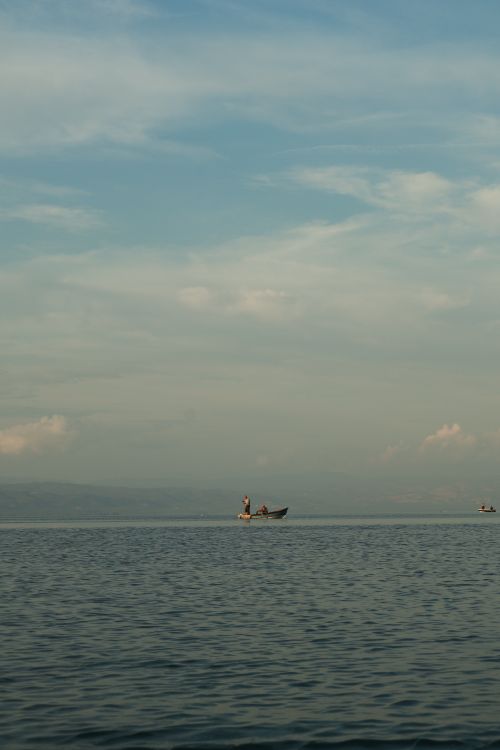 Free A lone fisherman in a boat on a calm lake under a blue sky, evoking peace and solitude. Stock Photo