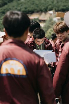 Men in maroon shirts gather outside, engaged in reading papers, with a scenic background.