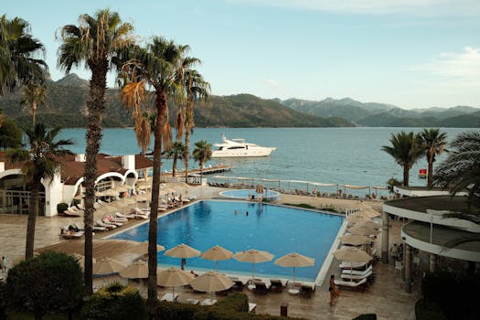 Elegant resort pool with ocean view, palm trees, and yacht docking. Ideal vacation spot.