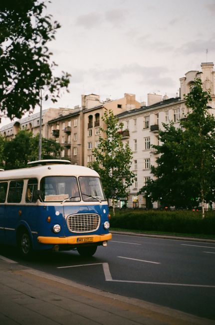 A retro blue and white bus on a Warsaw street with old buildings in the background.