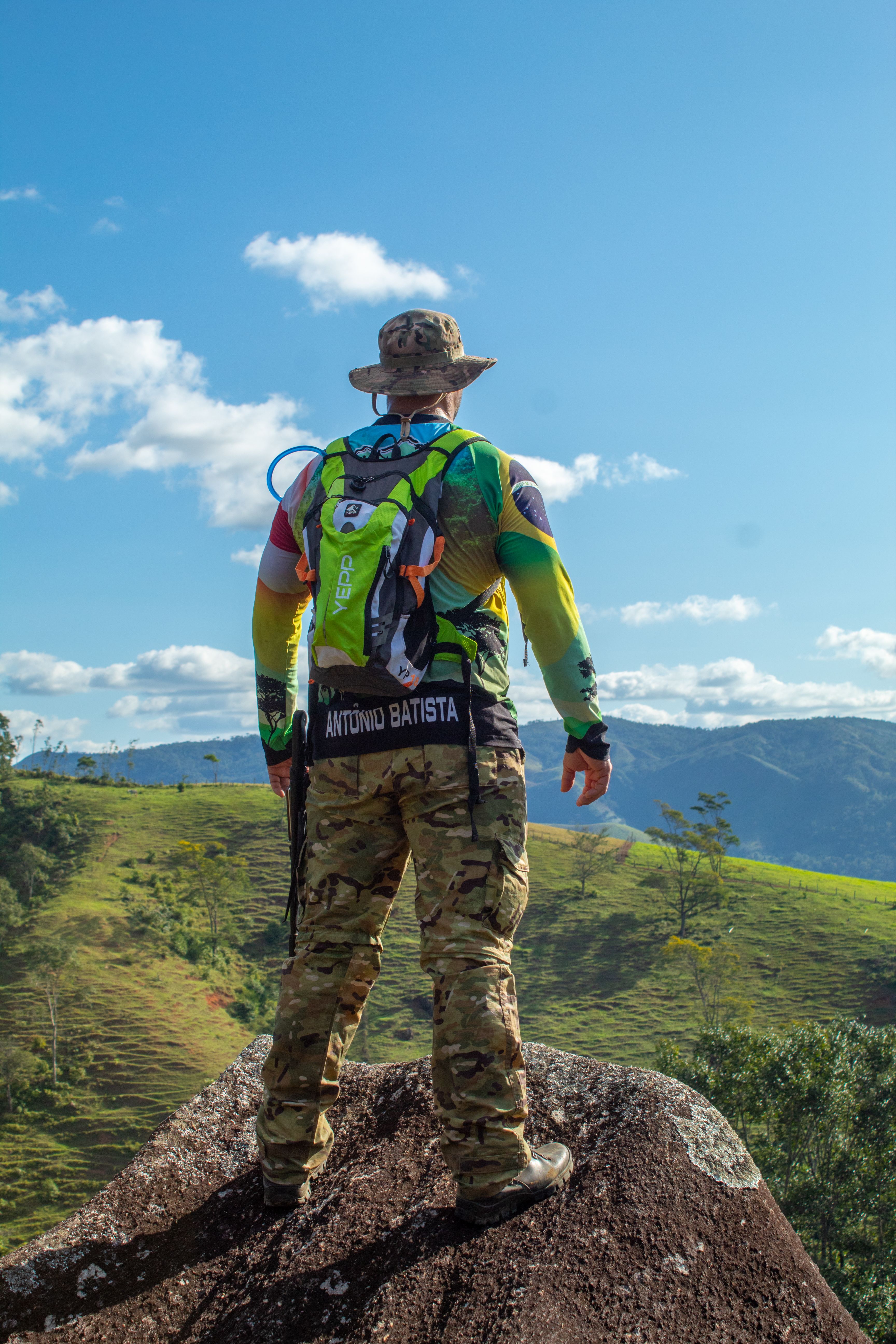 A hiker in camo attire and backpack overlooks a scenic mountain landscape on a sunny day.