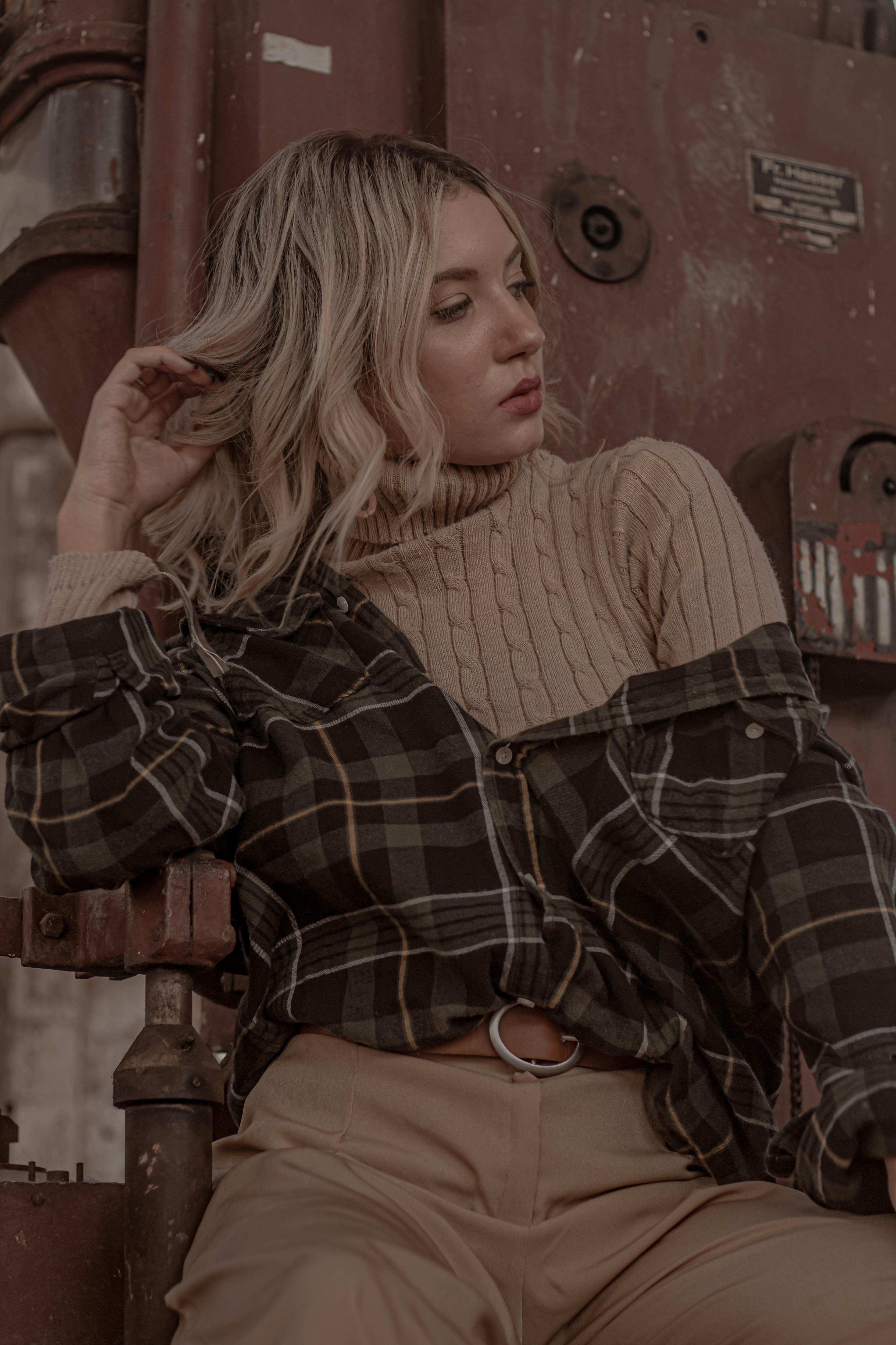 一位身穿格纹针织衫的金发女子在工业环境中摆出姿势，显得休闲而优雅