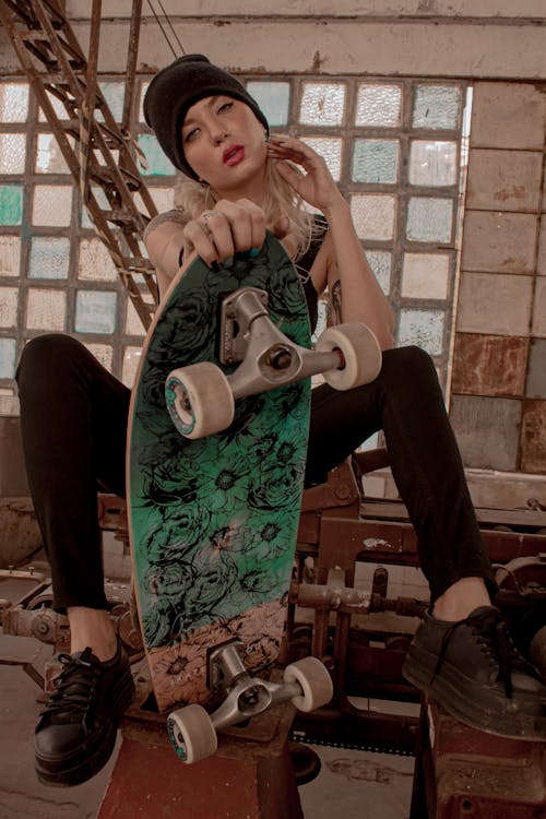 Photo of Sitting Woman in Black Outfit and Black Beanie Posing While Holding Skateboard