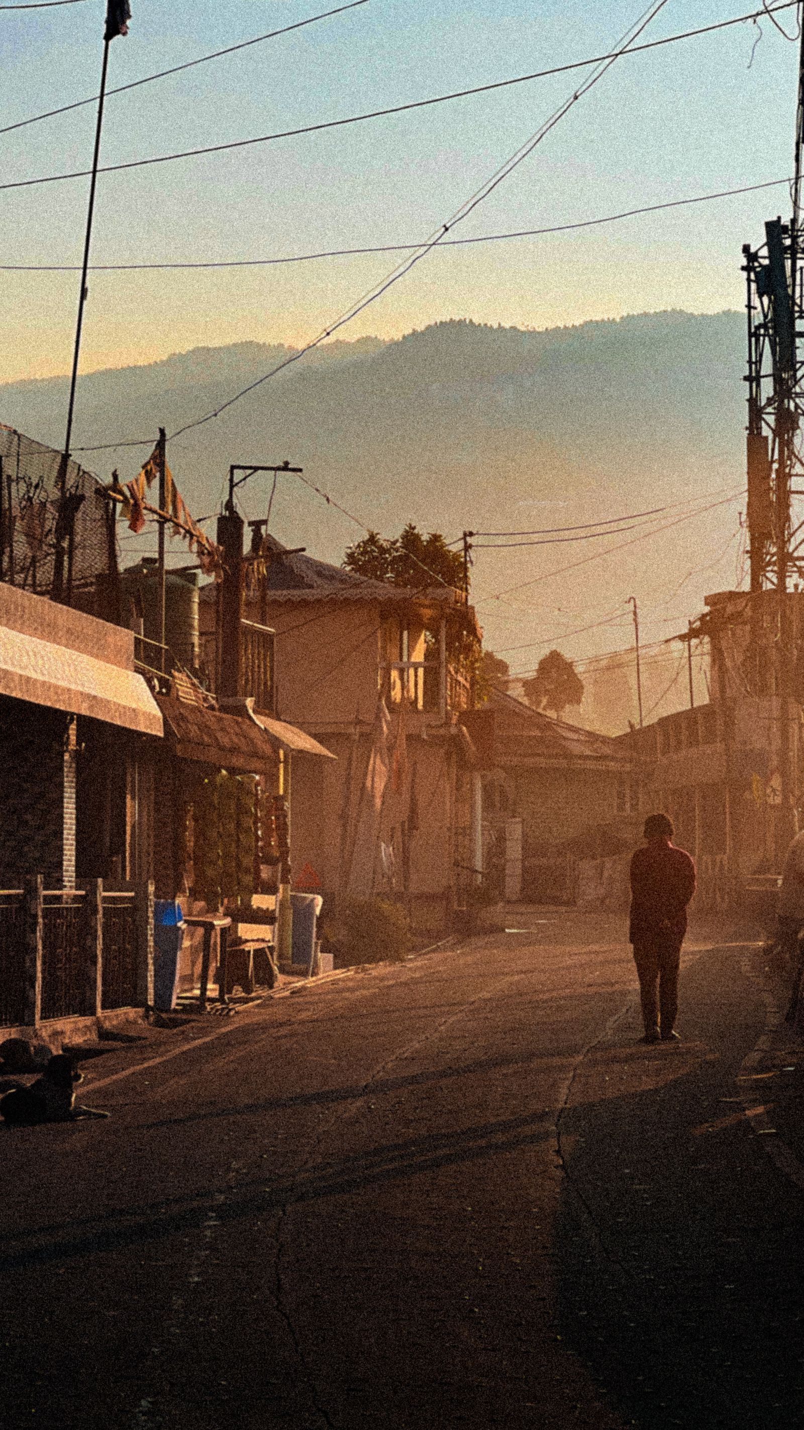A lone walker on an empty Darjeeling street at sunrise, shuttered shopfronts and hazy ridges glowing behind