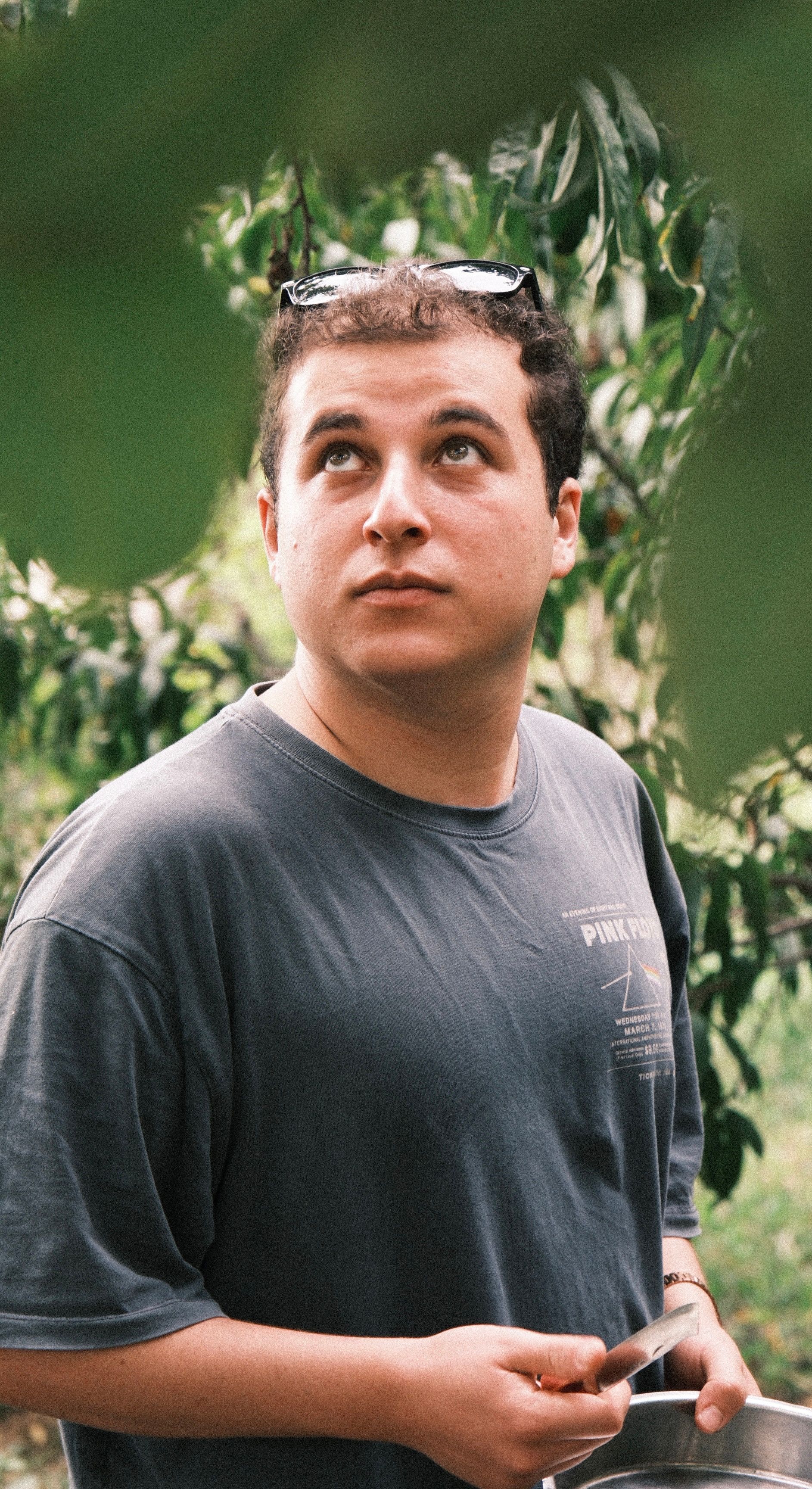 A young man holding a tool looks contemplatively upward surrounded by greenery.