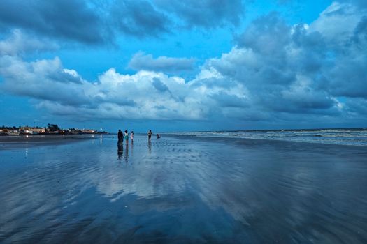 Dramatic cloudy sky reflected on the serene Mandarmani beach in India with people enjoying the scene.