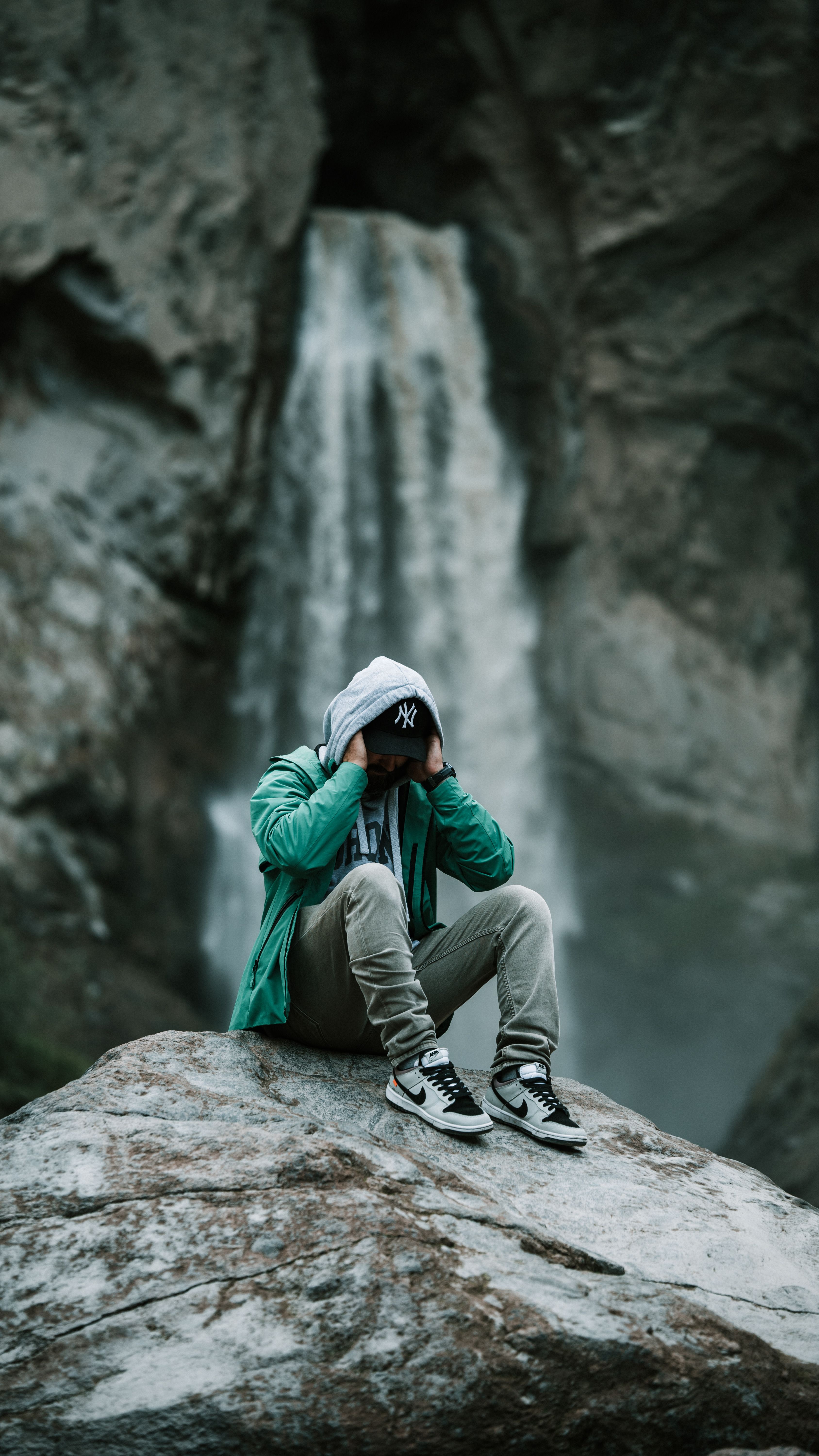 Man in green jacket sitting on rock with waterfall background, serene and contemplative atmosphere.