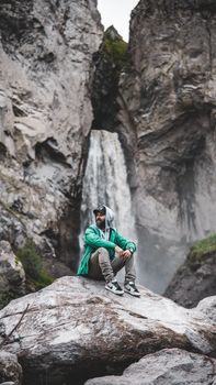 Explore tranquility and adventure with a man resting by a majestic waterfall in a mountainous setting.