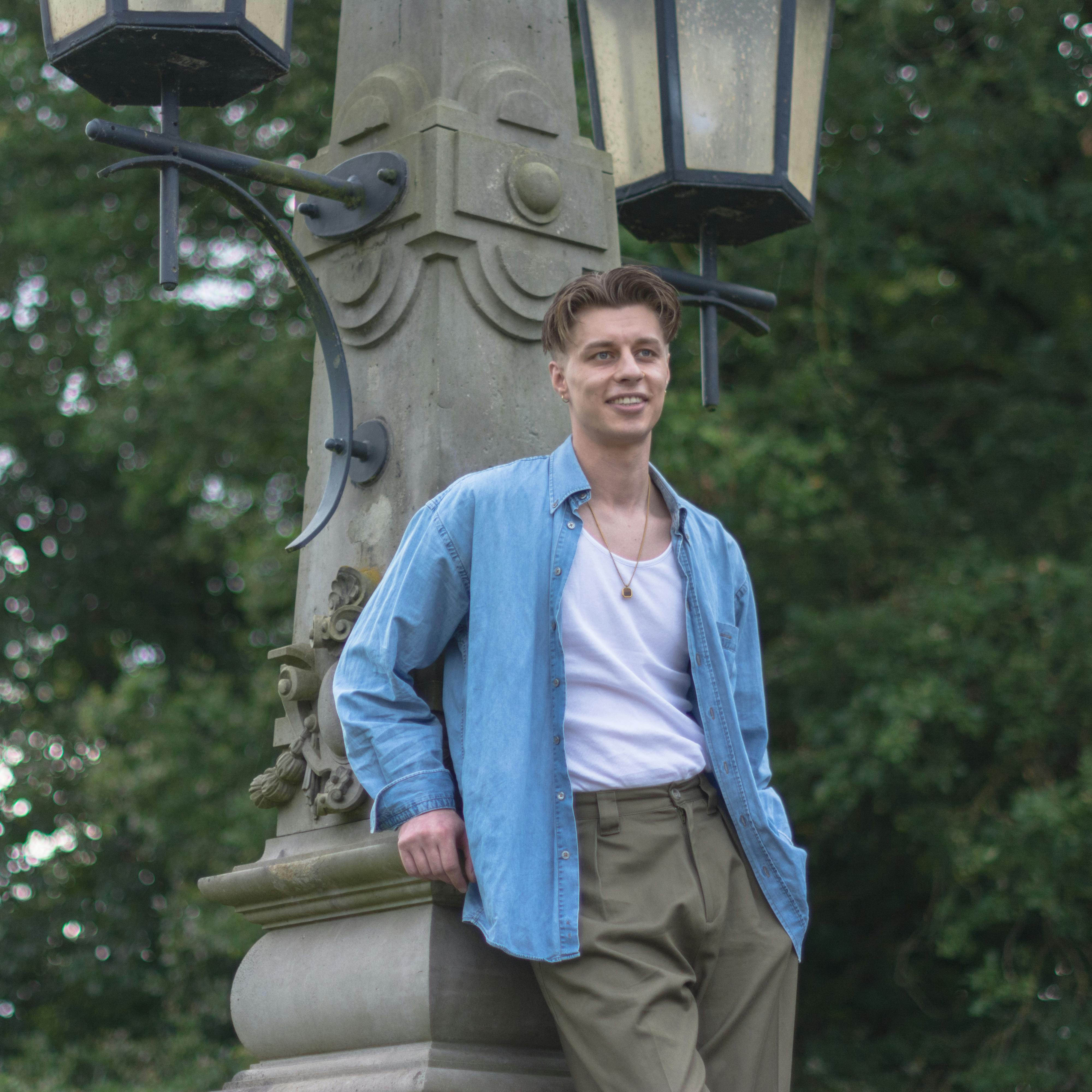 Stylish young man leans on a vintage street lamp in a lush outdoor setting.