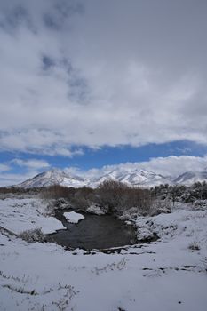 A peaceful snowy mountain scene with a calm stream and overcast sky.