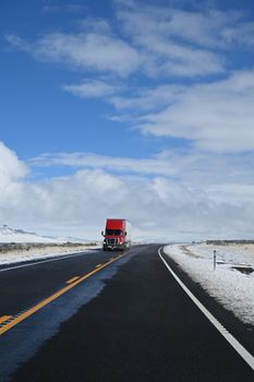 A red truck travels on a snowy, curved highway under a bright blue sky with clouds.