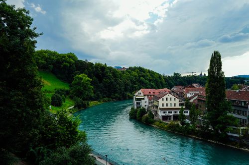 Free Picturesque view of a river winding through a town with lush green hills and dramatic clouds. Stock Photo