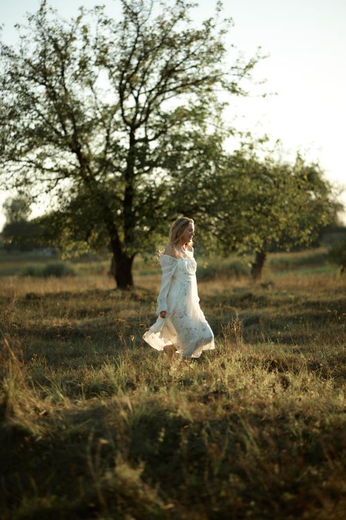 Free A woman in a white dress strolls through a sunlit meadow at golden hour. Stock Photo