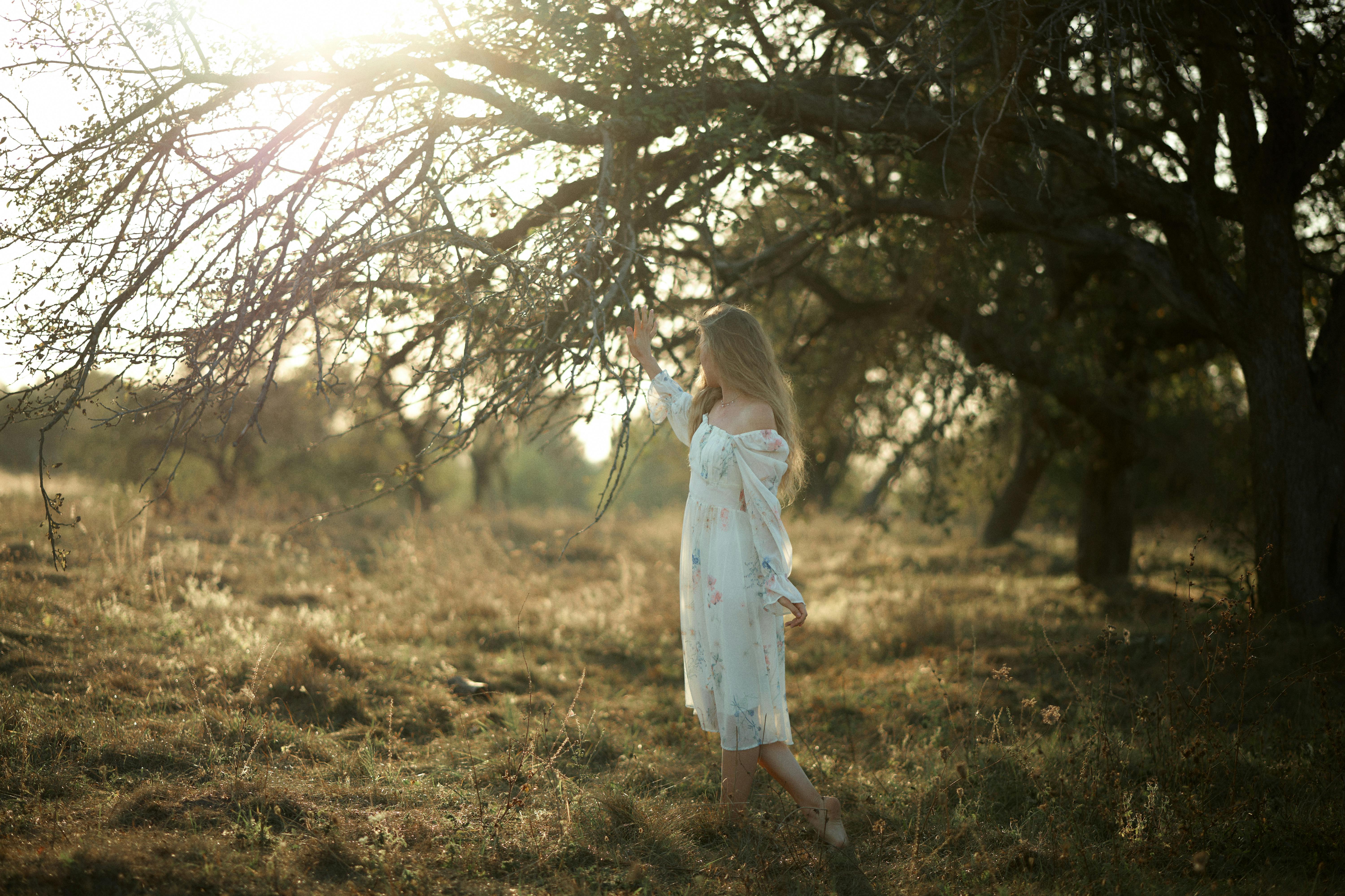 Woman in white dress enjoying sunlight in meadow under a tree.