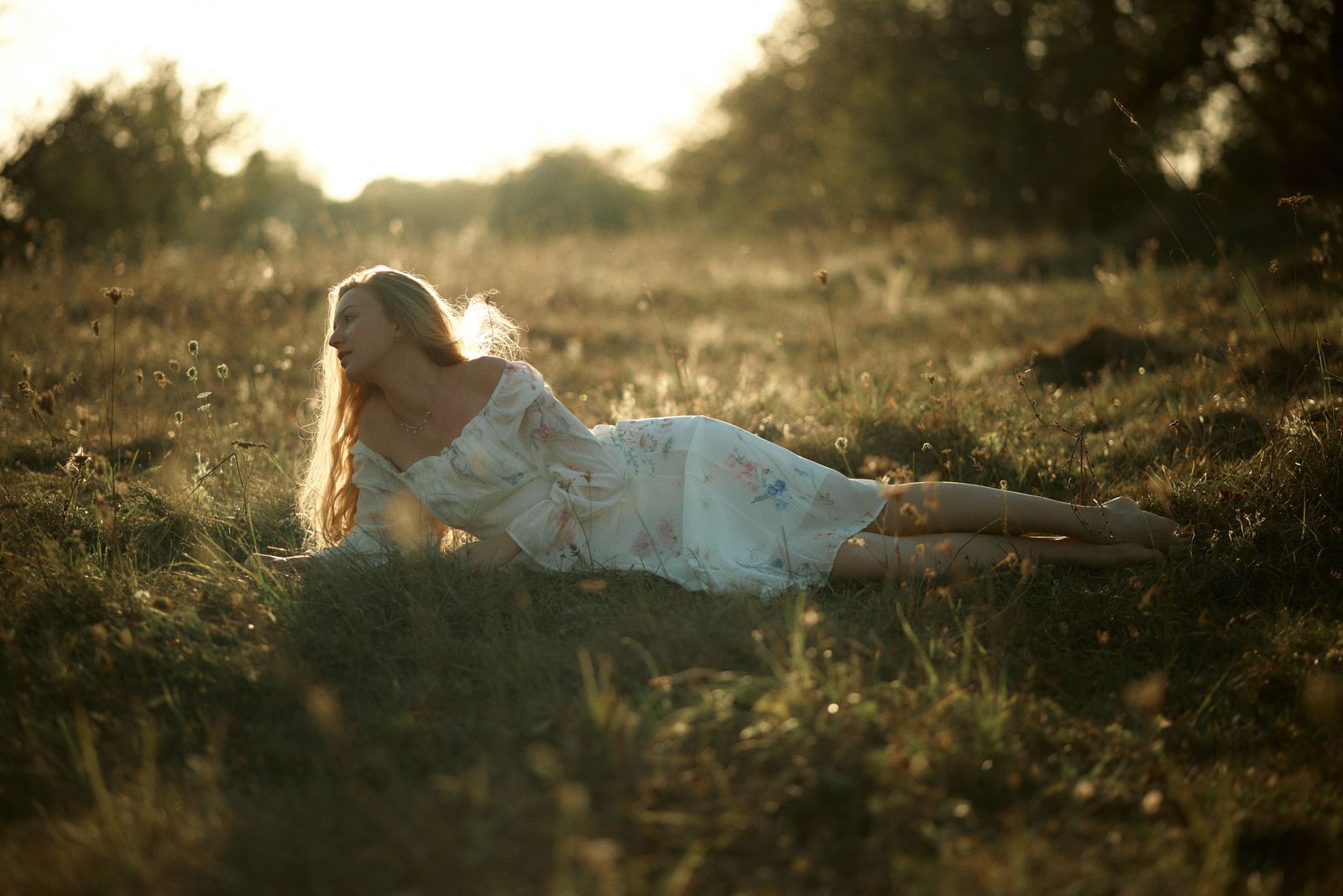 Woman in a white floral dress lying in a grassy meadow, backlit by warm golden sunlight