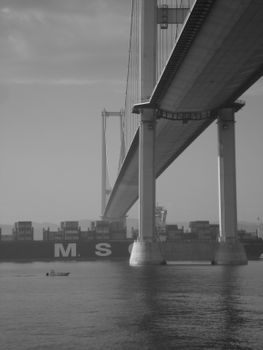 Black and white photo of a suspension bridge over a waterway with a large container ship underneath.
