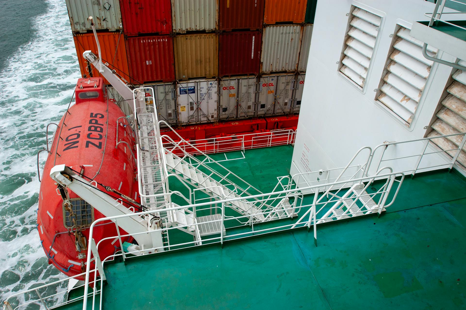 View of a container ship's deck featuring a red lifeboat and shipping containers.
