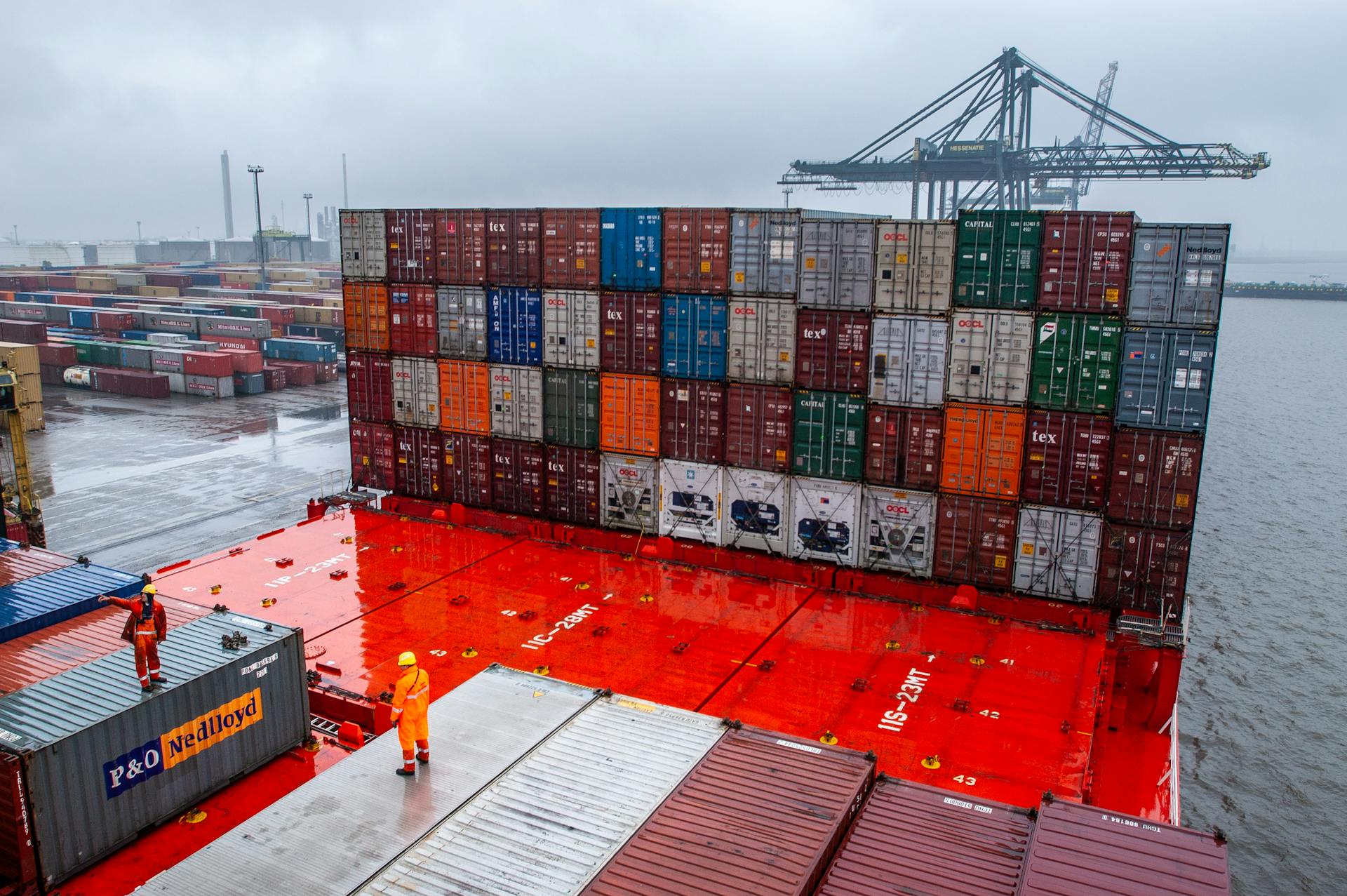 A busy container terminal with dock workers and shipping containers during rainy weather.