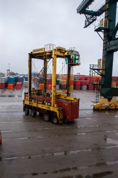 Yellow straddle carrier moving containers at a busy, rainy shipping port.