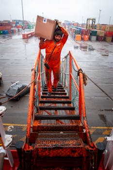 A maritime worker in orange coveralls carries a box up a ship's gangway in rainy weather at a port.