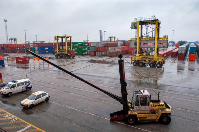 Container terminal with straddle carriers and stacked shipping containers under a cloudy sky.