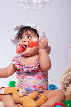 Cute baby wearing a summer outfit and hat holding red sunglasses, surrounded by toys.
