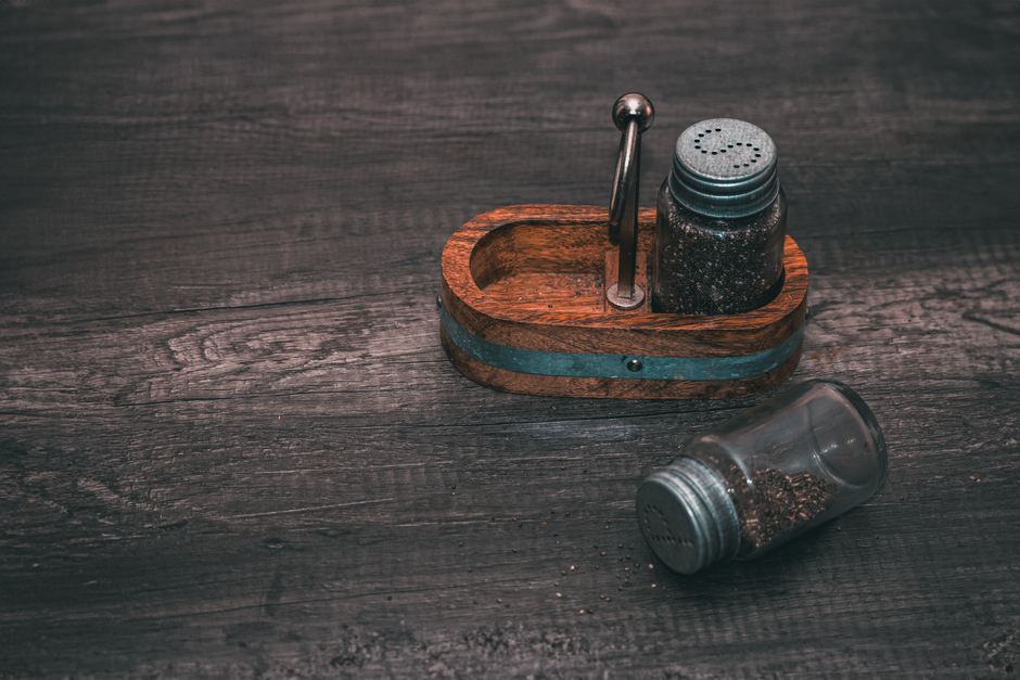 Dark-skinned Nigerian woman in a modest Lagos kitchen placing small sealed glass jars of dried iru into a wooden shelf beside other Nigerian condiments, natural window light, photorealistic